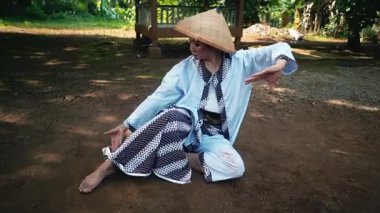 Geleneksel giysiler ve hasır şapka giyen bir insan yeşil ve arka planda bir çardak ile çevrili sakin bir açık hava ortamında yere oturur..