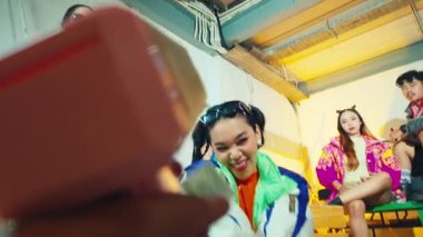 Canlı bir sahne. Renkli kıyafetler içindeki bir grup genç birlikte eğlenceli bir anın tadını çıkarıyor. Bir kişi pembe bir nesne tutuyor, muhtemelen bir oyuncak ya da kamera, diğerleri ise arka planda neşeli bir şekilde poz veriyor..