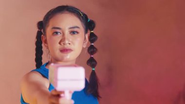 Saçları örgülü genç bir kadın pembe oyuncak bir silahı kameraya doğrultuyor, neşeli ve canlı bir atmosfer yaratıyor. Arka plan hafifçe bulanık, ifadesini ve oyuncağını vurguluyor..