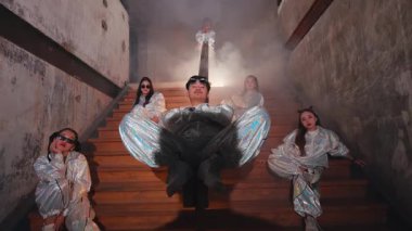 Mavi ışık ve dumanla aydınlatılmış, bir grup parlak kıyafetli sanatçıyı konu alan dramatik bir sahne. Bir sanatçı ön planda belirgin bir şekilde otururken, diğerleri onun etrafında toplanarak gizemli bir atmosfer yaratıyor..