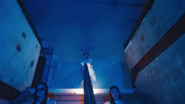 Merdivenler loş bir şekilde aydınlatılmış ve merdivenlerde oturan beyaz koruyucu elbiseli şahıslar var. Spot ışıklarıyla aydınlatılan ve dramatik bir atmosfer yaratan bir figür zirvede duruyor..