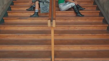 İki genç adam arka arkaya ahşap merdivenlerde oturuyor, biri kot ceketli, diğeri yeşil bluzlu ve gümüş pantolonlu, şık bir kontrast sergiliyorlar..