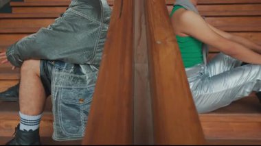 İki genç adam arka arkaya ahşap merdivenlerde oturuyor, şık ve rahat bir hava sergiliyorlar. Biri güneş gözlüğü ve kot ceket giyerken, diğerinin kolsuz bluzu ve kendinden emin bir ifadesi var..