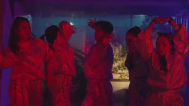 Parlak, yansıtıcı giysiler içinde bir grup dansçı canlı kırmızı ve mor ışık altında gösteri yapıyorlar. Atmosfer enerjik ve dinamiktir. Hareketleri ve ifadeleri gösterir..