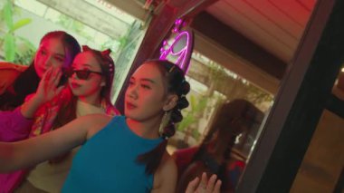 Canlı, neon ışıkları olan renkli bir ortamda poz veren üç genç kadın. Biri güneş gözlüğü takarken, diğeri neşeli bir ifadeye sahiptir. Arka planda yeşillik ve modern estetik yer alıyor..