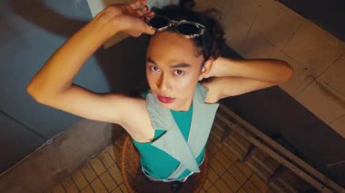 Şık bir birey, eşsiz bir kıyafetle, güneş gözlüğü ve metalik pantolonla, fayanslı duvarları olan endüstriyel bir ortamda dramatik bir şekilde poz verir..