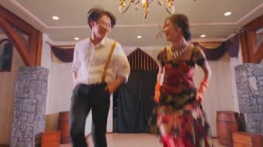 Güzel dekore edilmiş iç mekanda avizeyle dans eden neşeli bir çift. Kadın renkli bir elbise giyer, erkek ise günlük beyaz gömlek giyer. Ortam sıcak ve davetkar..