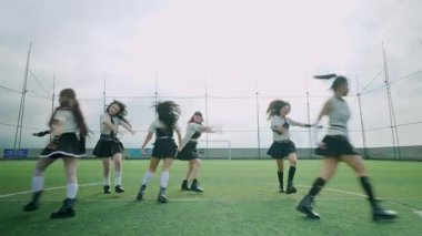 Bir grup dansçı bulutlu bir gökyüzünün altında bir futbol sahasında gösteri yapıyorlar. Etek ve şapkalar dahil şık kıyafetler giyiyorlar, senkronize hareketler sergiliyorlar..