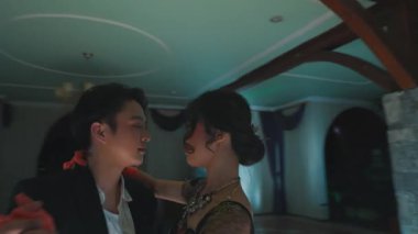 Loş bir balo salonunda zarif bir şekilde dans eden bir çift. Kadın turuncu eldivenli canlı kırmızı bir elbise giyerken erkek siyah bir takım elbise giyer. Atmosfer romantik ve sofistike..