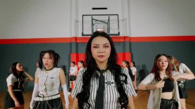 Dinamik bir dans performansı, şık kıyafetler içindeki bir grup genç kadının arka planda basketbol potası olan bir spor salonu ortamında enerjik koreografi sergilemesi..