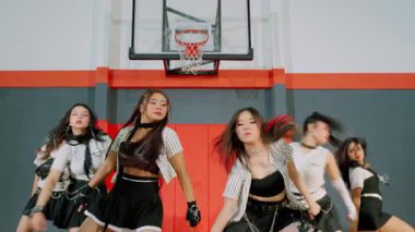 Jimnastik salonundaki beş kadın dansçıdan oluşan dinamik bir dans gösterisi. Zincirli şık kıyafetler giyiyorlar ve basketbol potası önünde senkronize hareketler yapıyorlar..