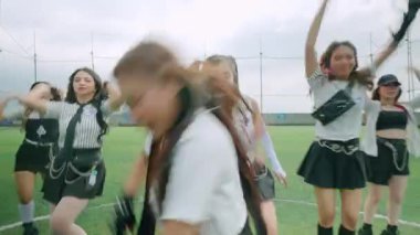 Bir grup dansçı yeşil bir çim sahasında, şık siyah beyaz kıyafetler giyerek gösteri yapıyorlar. Sahne dinamik hareketleri ve ifadeleri yakalıyor, koreografili bir rutini gösteriyor..