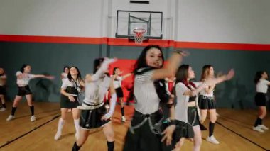 Bir spor salonundaki dinamik dans gösterisi, şık kıyafetler içindeki enerjik dansçılardan oluşan bir grup. Odak noktası, dalgalı saçlı, coşkulu ve hareketli merkezi bir dansçı..