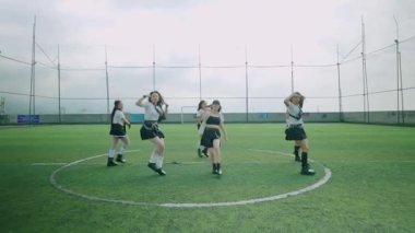 Altı dansçıdan oluşan bir grup yeşil bir çim sahasında, şık kıyafetler giymiş. Sahne, arka planda bulutlu bir gökyüzü ile dinamik bir hareket ve enerji yakalar..