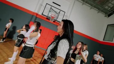 Bir grup dansçı spor salonunda, çizgili kıyafetler giyip senkronize bir gösteri yapıyorlar. Arka planda bir basketbol potası ve canlı bir spor ortamı yer alıyor..