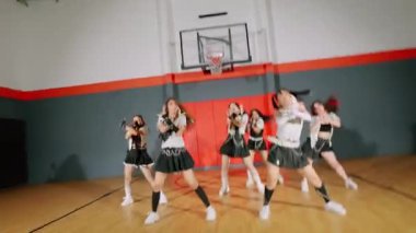 Bir spor salonundaki dinamik dans gösterisinde şık kıyafetler içinde birden fazla dansçı olacak. Arka planda bir basketbol potası ve renkli duvar panelleri var. Enerjik bir atmosfer yaratıyorlar..