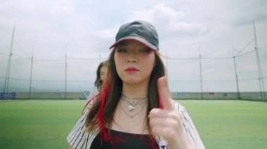 Genç bir kadının enerjik bir şekilde dans ettiği canlı bir spor sahnesi. Şapka ve çizgili bir gömlek giyiyor, neşeli bir ifadesi var. Arka planda, diğer dansçılar görülebilir, canlı bir atmosfer yaratıyorlar..