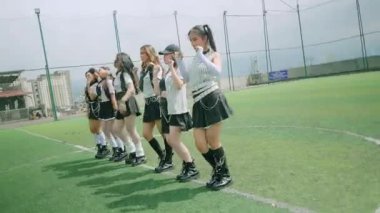 Spor sahasında dans eden, şık kıyafetler giyen bir grup genç kadının dinamik bir sahnesi. Arka planda bir spor tesisi ve bulutlu bir gökyüzü var. Enerjik bir atmosfer yaratıyor..