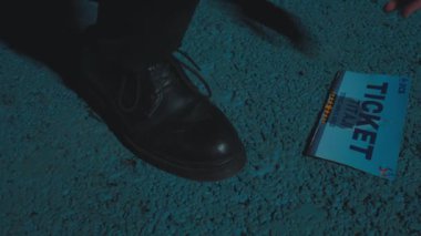 Yakınlarda bir biletle desenli bir yüzey üzerinde siyah bir ayakkabının yakın çekimi. Sahne mavi bir tonla aydınlatılıyor, dramatik bir atmosfer yaratıyor..