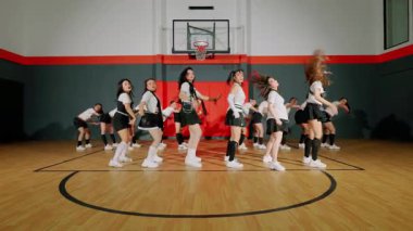 Spor salonunda dinamik bir dans gösterisi, koordine kıyafetlerle dans eden bir grup dansçı. Arka planda basketbol potası olan enerjik hareketler sergiliyorlar..