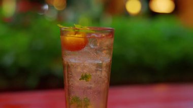 Uzun bardakta servis edilen ferahlatıcı bir kokteyl, kiraz ve nane yapraklarıyla süslenmiş, içkinin içinde kabarcıklar var. Arka plan hafifçe bulanık, sıcak bir ortam yaratıyor..