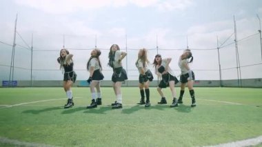 Altı dansçıdan oluşan bir grup, yeşil bir alanda, parlak bir gökyüzünün altında koreografi gösterisi yapıyorlar. Şık kıyafetler giyiyorlar, dinamik pozlar ve ifadeler sergiliyorlar..