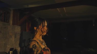 Zarif elbiseli bir kadın loş bir odada dikiliyor ve kıyafetinin detaylarını anlatıyor. Ortam sofistike, arka planda hafif ışıklandırma ve dekoratif öğeler var..