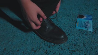 Siyah bir ayakkabıya ayakkabı bağcıkları bağlayan bir kişinin yakın plan resmi, ve yakınlarda dokulu bir yüzey üzerinde mavi bir bilet var..