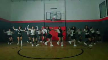 Bir grup dansçı bir basketbol salonunda koreografi gösterisi yapıyor. Aynı kıyafetleri giyiyorlar ve arka planda basketbol potası olan senkronize hareketler sergiliyorlar..