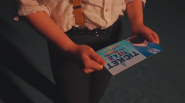 Elinde mavi arka planı olan, beyaz gömlek ve pantolon askısı takan biri. Bilette 'TICKET' kelimesi belirgin bir şekilde gösteriliyor, bir olay veya performans öneriliyor.