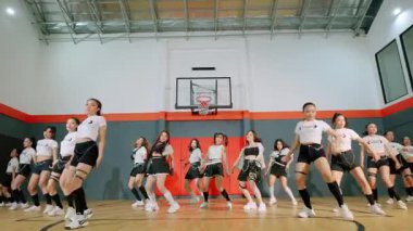 Bir grup dansçı bir basketbol salonunda koreografi gösterisi yapıyor. Dansçılar şık atletik kıyafetler giyip hareketlerinde bir enerji ve hassasiyet karışımı sergiliyorlar..
