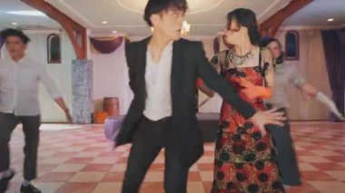 Bir grup dansçı güzel dekore edilmiş bir salonda gösteri yapıyorlar. Merkez çift zarif giyinmiş, renkli elbiseli kadın ve takım elbiseli adamla birlikte. Diğer dansçılar arka planda, canlı bir dans ortamı sergiliyorlar..