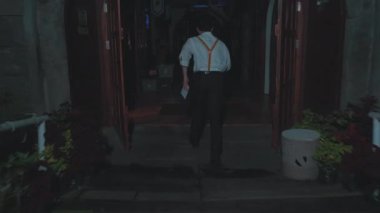 Loş bir odada beyaz bir gömlek, pantolon askısı ve siyah pantolon giyerek yürüyen biri. Arka planda dekoratif elementler ve hafif ışıklandırma var. Gizemli bir atmosfer yaratıyor..