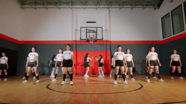 Bir grup dansçı jimnastik salonunda sahne alıyor, arka planda basketbol potası olan aynı kıyafetleri giyiyorlar. Koreografi enerjik ve senkronize, yeteneklerini sergiliyor..