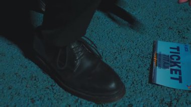 Yakından bakınca, mavi desenli bir yüzey üzerinde siyah deri bir ayakkabı ve yanında da bir bilet var. Ayakkabı cilalanmış ama aşınma izleri var ve bilette kalın bir metin var..