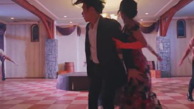 Resmi giyinmiş, ortasında el ele tutuşan bir çiftin olduğu canlı bir dans sahnesi. Kadın karmaşık tasarımları olan zarif bir kırmızı elbise giyer, erkek ise siyah takım elbise giyer. Arka planda, diğer dansçılar bir festival düzenliyorlar.