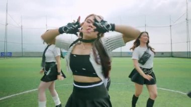 Yeşil çim sahasında gösteri yapan altı kadın dansçıdan oluşan bir grup. Şık kıyafetler giyip, bulutlu bir arkaplana karşı dinamik dans hareketleri sergiliyorlar..