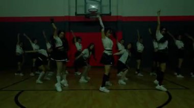 Bir spor salonundaki dinamik dans gösterisi. Bir grup dansçının eşgüdümlü kıyafetleri var. Orta atlama, enerji ve senkronizasyon sergiliyorlar. Arka planda basketbol potası var..
