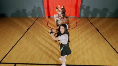 Bir grup genç dansçı bir spor salonunda koreografi gösterisi yapıyor. Eşleşen kıyafetler giyiyorlar, bir basketbol sahasının önünde enerjik hareketler sergiliyorlar..