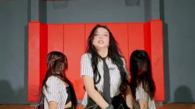 Dinamik bir dans gösterisi. Çizgili kıyafetli bir kadın dansçı. Etrafında iki dansçı daha var. Arka plan kırmızı duvarları ve basketbol potası olan bir spor salonu..