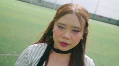 Uzun saçlı ve makyajlı, çizgili bir gömlek giyen genç bir kadın, parlak güneş ışığı altında bir futbol sahasında neşe içinde poz veriyor. Arkaplanda yeşil çim sahası ve kale direkleri yer alıyor.