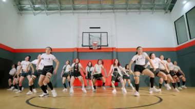 Bir grup genç kadın spor salonunda senkronize dans gösterisi yapıyor. Sportif kıyafetler giyiyorlar, enerji ve takım çalışması sergiliyorlar. Arka plandaki basketbol potası atletik atmosfere katkı sağlıyor..