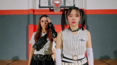 Basketbol sahasında kendine güvenen iki genç kadın, 'TOXIC' sözcüğüyle uyumlu çizgili kıyafetler giyiyorlar. Arka planda basketbol potası ve renkli duvarlar var..