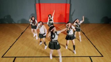 Altı dansçıdan oluşan bir grup bir spor salonunda koreografi gösterisi yapıyor. Şık kıyafetler giyiyorlar, kırmızı bir duvar ve ahşap zemine karşı enerjik hareketler sergiliyorlar..