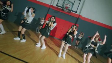 Bir grup dansçı bir spor salonunda koreografi gösterisi yapıyor. Dansçılar uyumlu kıyafetler giyer, senkronize hareketler sergilerler. Basketbol potası ve sahası arka planda görülebiliyor ve atletik atmosfere ekleniyor..