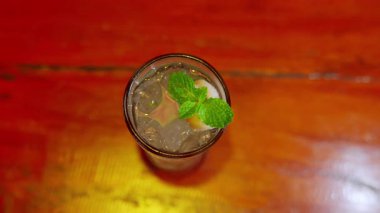 Buzlu ferahlatıcı bir içecek ve üstünde bir tutam nane, uzun bir bardakta servis ediliyor. Arkaplanda ahşap bir masa yüzeyi var.