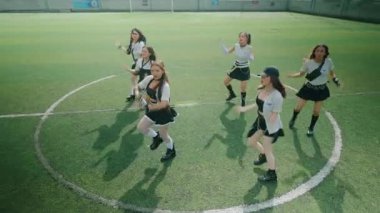 Futbol sahasında dans eden altı genç kadından oluşan bir grup. Sportif kıyafetler giyiyorlar, enerjik hareketler sergiliyorlar. Arka planda yeşil çim ve kale direkleri görünüyor..