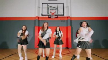 Spor salonunda bir dansçı, şık bir kıyafet, çizgili bir bluz ve etek giyiyor. Arka planda bir basketbol potası ve kırmızı duvarlar var, canlı bir atmosfer yaratıyor..