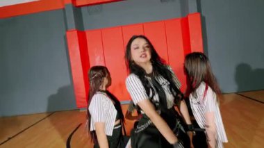 Basketbol salonunda dans eden beş genç kadından oluşan bir grup. Sportif kıyafetler giyiyorlar, etek ve şort karışımı, enerjik hareketler sergiliyorlar. Spor salonunda bir basketbol potası ve canlı bir renk düzeni var..