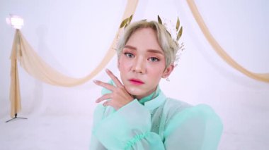 Bir model zarif bir şekilde nane yeşili elbisenin içinde saçlarında çiçek aksesuarlarıyla poz verir. Arka plan örtülü kumaş içeriyor, sakin bir atmosfer yaratıyor..